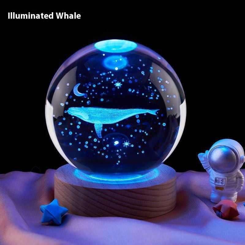 Glass ball with whale design on a wooden base, illuminated by blue light, with an astronaut figure in the background.