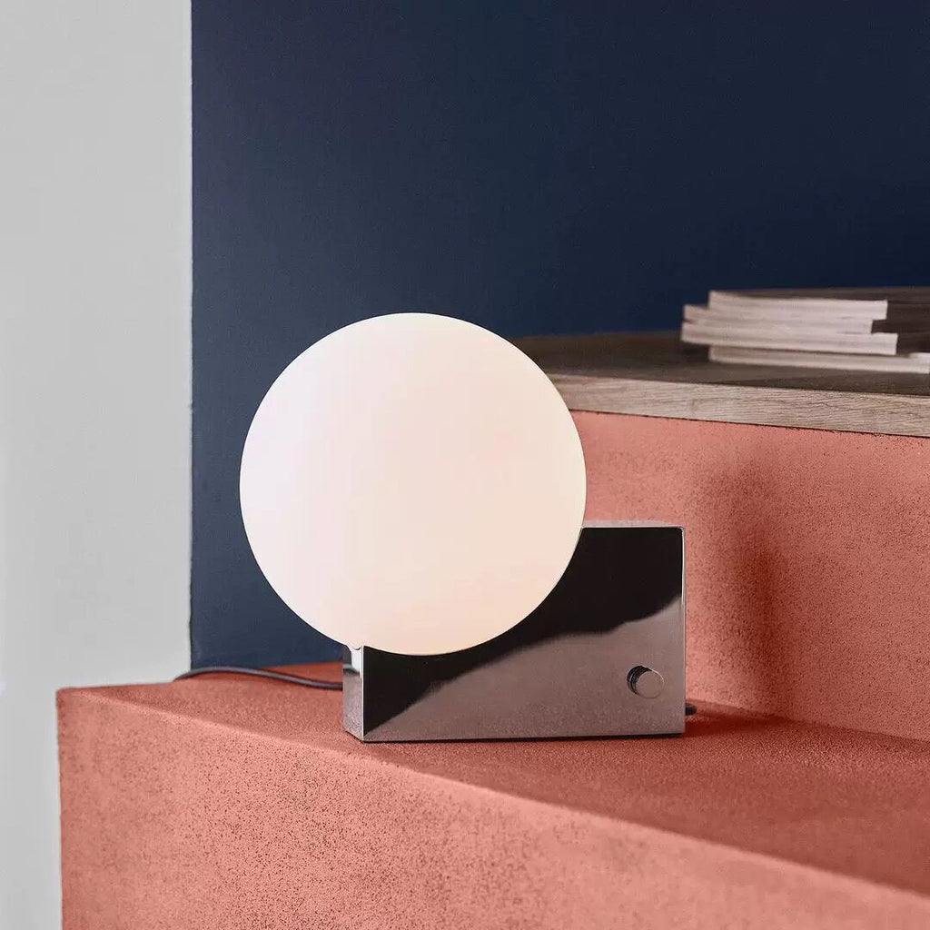 Modern table lamp with a spherical white shade on a pink platform against a blue wall.
