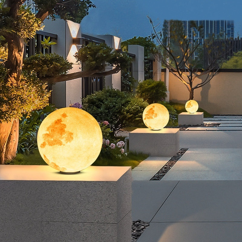Decorative moon-shaped lights on stone plinths in a garden setting with modern buildings in the background.