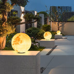 Decorative moon-shaped lights on stone plinths in a garden setting with modern buildings in the background.